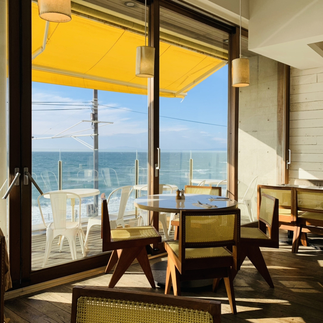 bills七里ガ浜内観であり、そこにはテーブルと椅子が数セットあり、窓の外には広がる綺麗な海岸と地平線が見えます。