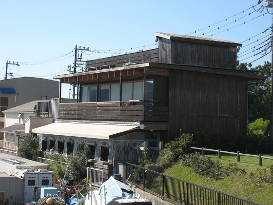 江ノ島小屋の外観であり、おしゃれな雰囲気の木造建物と雲ひとつない青空が見えます。